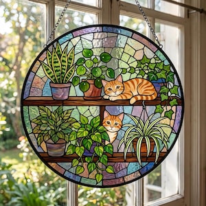May include: A circular stained glass suncatcher featuring two orange tabby cats resting among various potted houseplants on two wooden shelves. The background consists of a mosaic of multicoloured glass pieces, all framed in black metal with a hanging chain.