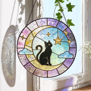 May include: A circular stained glass suncatcher featuring a black cat silhouette sitting on a yellow crescent moon. The background displays a gradient of blue and purple glass with white clouds and several yellow stars. It hangs from a metal chain.