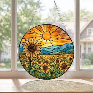 May include: A circular stained glass suncatcher featuring a vibrant sunflower field under a bright yellow sun and blue mountain range. The piece is framed in black metal and hangs from a thin black chain, casting colourful light reflections.