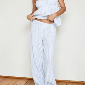 Puede incluir: Un conjunto de pijama a rayas azul claro y blanco. El conjunto incluye una camiseta sin mangas con tirantes finos y un busto fruncido, y pantalones de pernera ancha. La tela parece ligera. El modelo lleva zapatos marrones.