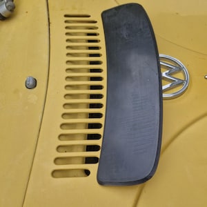 Può includere: Un componente in gomma nera è appoggiato sul cofano giallo di un'auto d'epoca. Il cofano presenta una serie di fessure parallele e un'emblema cromato. La vernice dell'auto mostra segni di usura.