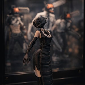 Könnte beinhalten: Schwarze Figur einer Frau in einem Kleid, mit einem maskenartigen Kopf und langen Handschuhen. Weitere ähnliche Figuren sind im Hintergrund hinter Glas zu sehen. Das Kleid hat ein strukturiertes, geschichtetes Design.