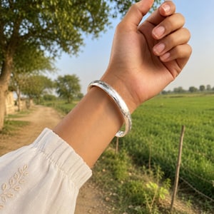 Puede incluir: Una pulsera rígida de color plateado con una superficie lisa, pulida y ligeramente irregular. La pulsera se lleva en la muñeca y presenta un perfil redondeado con un acabado metálico reflectante que capta la luz natural.