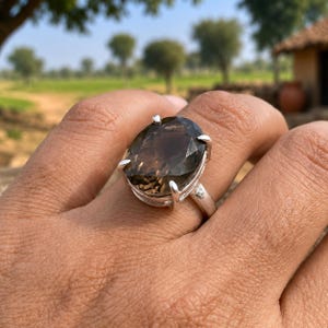 May include: A silver ring featuring a large, oval-cut, translucent brown smoky quartz gemstone. The stone is secured in a prong setting with four metal claws, mounted on a polished silver band that encircles the finger.