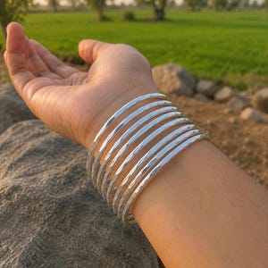 Op de afbeelding: Een set van acht individuele zilverkleurige armbanden die samen om de pols worden gedragen. Elke armband heeft een glad, afgerond profiel met een gepolijst, reflecterend oppervlak dat het licht opvangt en een gestapeld uiterlijk op de arm creëert.