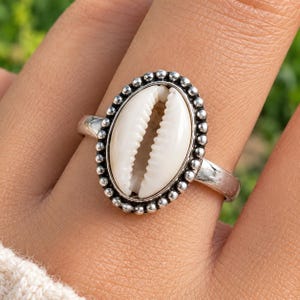 May include: A silver ring featuring a central oval cowrie shell set within a beaded metal border. The band is smooth and polished, framing the white, ridged shell which is positioned vertically as the focal point of the jewellery piece.