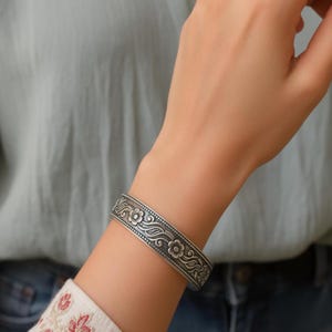 May include: A silver-toned cuff bracelet featuring an embossed floral pattern with swirling vine details. The metal surface has an oxidized finish that highlights the intricate engraved flowers and leaves along the entire length of the band.