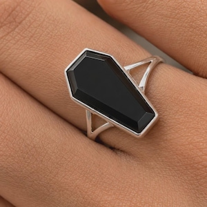 May include: A silver ring featuring a faceted black stone cut into a coffin shape. The ring has a split shank design that connects to the sides of the stone setting, creating a geometric look on the finger.
