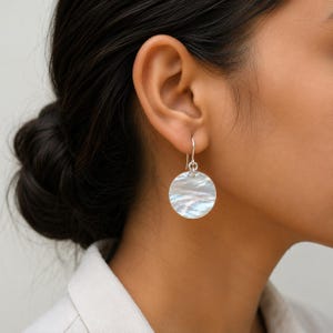 Può includere: Un orecchino circolare con un disco in madreperla iridescente lucida, sospeso a un semplice gancio in argento. Il disco presenta una superficie liscia e ondulata con sottili tonalità bianche e perlacee che catturano la luce.
