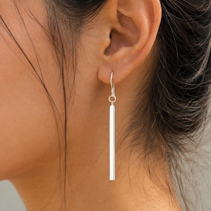 Peut inclure: Une boucle d'oreille argentée composée d'un long pendentif en forme de barre rectangulaire fine, suspendu à un crochet simple. La surface métallique est lisse et réfléchissante, tombant verticalement depuis le lobe de l'oreille.