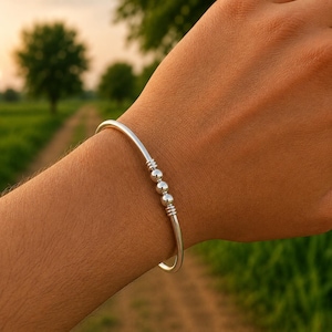 May include: A polished silver-tone bangle bracelet featuring a smooth, circular band. The centre of the bracelet is adorned with three small, spherical silver beads flanked by two sets of three thin, horizontal engraved lines on the metal surface.