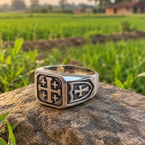May include: A silver ring with a 925 hallmark stamped on the inner band. The face of the ring features a square Jerusalem cross design with a dark, recessed background. The side of the ring displays a shield emblem containing a smaller cross.