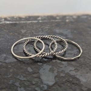 May include: A set of three silver-toned rings. One ring features a smooth, thin band, another has a twisted rope-like texture, and the third ring has a subtle, hammered or faceted surface finish. The rings are arranged in an overlapping cluster.