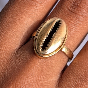 Può includere: Un anello color oro caratterizzato da un elemento centrale prominente a forma di conchiglia di ciprea ovale. La conchiglia presenta una finitura metallica liscia con un'apertura centrale scura e frastagliata che corre verticalmente.