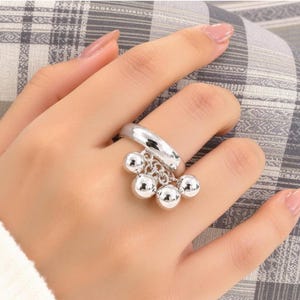 May include: A polished silver-tone ring featuring a smooth, solid band. Attached to the front of the band are four small, round, reflective silver-tone spheres that hang from delicate individual chain links, creating a dangling charm effect.