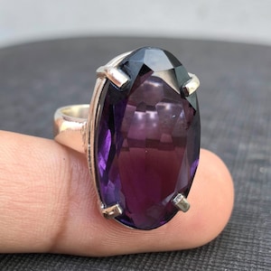 May include: A silver ring featuring a large, oval amethyst gemstone. The deep purple stone is held in place by four prongs. The ring is displayed on a finger, with the facets of the stone reflecting light.
