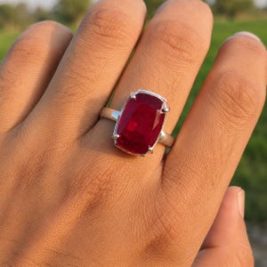May include: A silver ring featuring a large, rectangular, faceted deep red gemstone set in a four-prong setting. The stone has a cushion-cut appearance with visible internal clarity and light reflection across its polished surface.