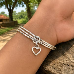 May include: A silver-toned bangle bracelet consisting of three stacked bands with a hammered, textured finish. A small, open-heart-shaped charm hangs from a circular ring that connects the three bands together, creating a layered appearance.