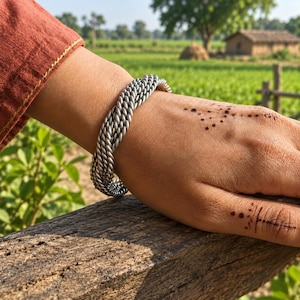 Può includere: Un bracciale a fascia color argento caratterizzato da un design a corda intrecciata a più fili. Il bracciale è composto da diversi fili metallici intrecciati insieme per creare un aspetto testurizzato e intrecciato attorno al polso.