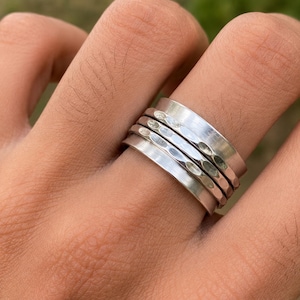Puede incluir: Un anillo ancho de color plateado con un diseño de varias bandas. El anillo consta de dos bandas exteriores lisas y dos bandas centrales con un acabado martillado y texturizado. La superficie metálica tiene un brillo pulido y un tono plateado uniforme.