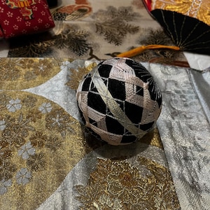 Puede incluir: Una bola Temari negra y dorada con un patrón geométrico, sobre una tela de brocado dorada y plateada. La bola está intrincadamente cosida con hilo metálico. Otros textiles japoneses están en el fondo.
