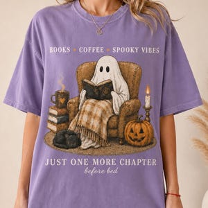 Op de afbeelding: Een paars t-shirt met een afbeelding van een geest die in een fauteuil zit en een boek leest. De print bevat een stapel boeken, een koffiemok, een pompoen, een zwarte kat en de tekst: BOOKS COFFEE SPOOKY VIBES JUST ONE MORE CHAPTER before bed.