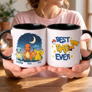May include: Two white ceramic mugs with black handles and rims. One mug features a night scene with cartoon characters and names. The other mug says "Best Dad Ever" with a lightning bolt and stars. The mugs are held by a person.