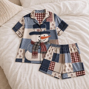 May include: A short-sleeve button-down pajama shirt and matching shorts set featuring a patchwork design of blue, white, and red plaid and snowflake patterns. The shirt displays a large central graphic of a snowman wearing a hat and scarf.