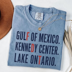 Op de afbeelding: Een blauw T-shirt met korte mouwen met de tekst GULF OF MEXICO, KENNEDY CENTER en LAKE ONTARIO in donkerblauw gedrukt, waarbij de letters F, D en T in het rood zijn gemarkeerd. Het T-shirt heeft een Comfort Colors merklabel in maat S.