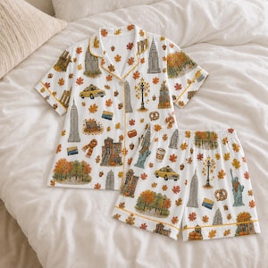 Op de afbeelding: Een witte pyjamaset met een pyjamashirt met korte mouwen en knoopsluiting en een bijpassende short, voorzien van een herhalende print van New Yorkse bezienswaardigheden, gele taxi's, herfstbladeren, pretzels en koffiebekers, afgewerkt met gele biesjes.