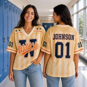 Puede incluir: Una camiseta de cuello en V con rayas amarillas y blancas, con el texto WildCats y la letra W en el frente, y el nombre JOHNSON junto al número 01 en la espalda. Las mangas muestran el número 01, rayas naranjas y estrellas azules en los hombros.