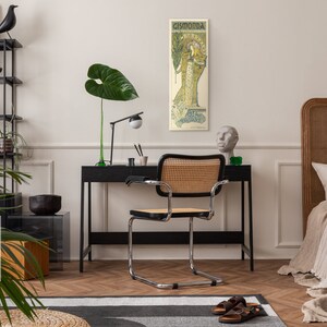 May include: A black and white poster with the text "Cismonda" in a decorative font. The poster features a woman in a long, flowing dress with a floral design. The poster is hanging on a white wall in a room with a black desk, a chair, and a bed.
