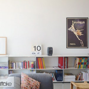 May include: A white bookshelf with books, games, and a calendar that says "10 SUN IX" on it. There is a framed poster on the wall that says "Ph&eacute;bus Paris" with a woman in a vintage dress riding a bicycle.