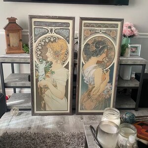 May include: Two framed art prints with a vintage aesthetic. Each print features a woman in a flowing gown with intricate floral designs and Art Nouveau style details. The prints are in brown wood frames.
