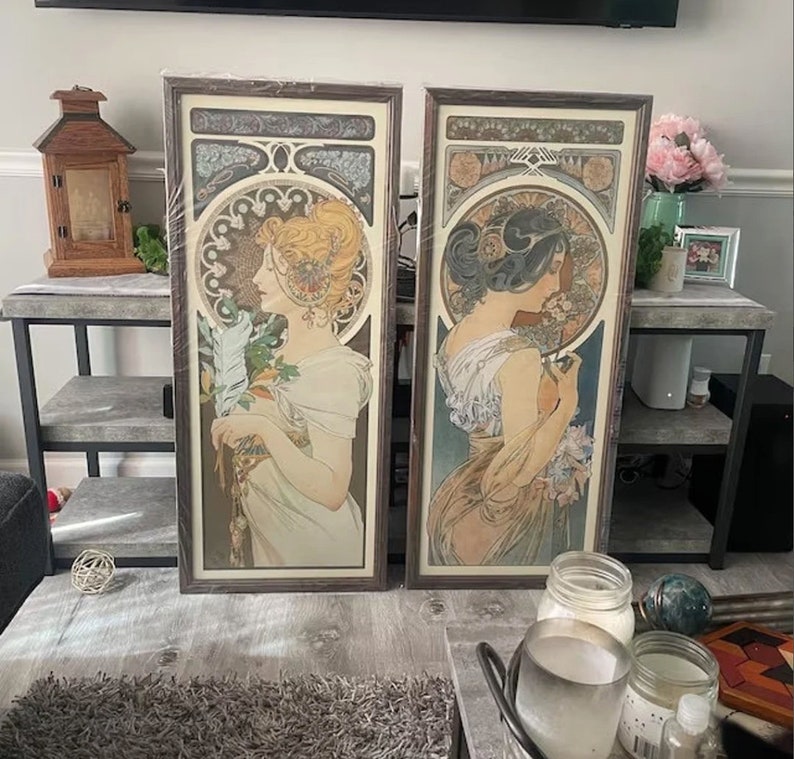 May include: Two framed art prints with a vintage aesthetic. Each print features a woman in a flowing gown with intricate floral designs and Art Nouveau style details. The prints are in brown wood frames.