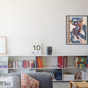 May include: A framed vintage poster with the word "PROGRAMME" vertically on the left side. The poster features a nude figure intertwined with blue and green paisley-patterned serpents. The poster is displayed above a white shelf with books and decorative items.