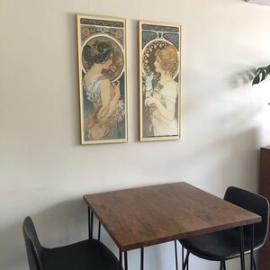 May include: Two framed art prints of women in Art Nouveau style, hanging on a white wall above a wooden table with black chairs.