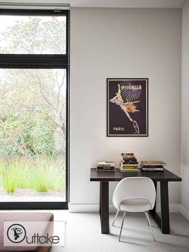 May include: A white desk chair with a black metal frame sits in front of a dark wood desk with a stack of books and a framed vintage poster that reads "Phoebus Paris".