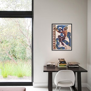May include: A framed art print featuring a nude figure with blue and white serpentine forms. The artwork is displayed on a white wall above a dark wood desk with books and a white chair. The room has a large window with a view of greenery.