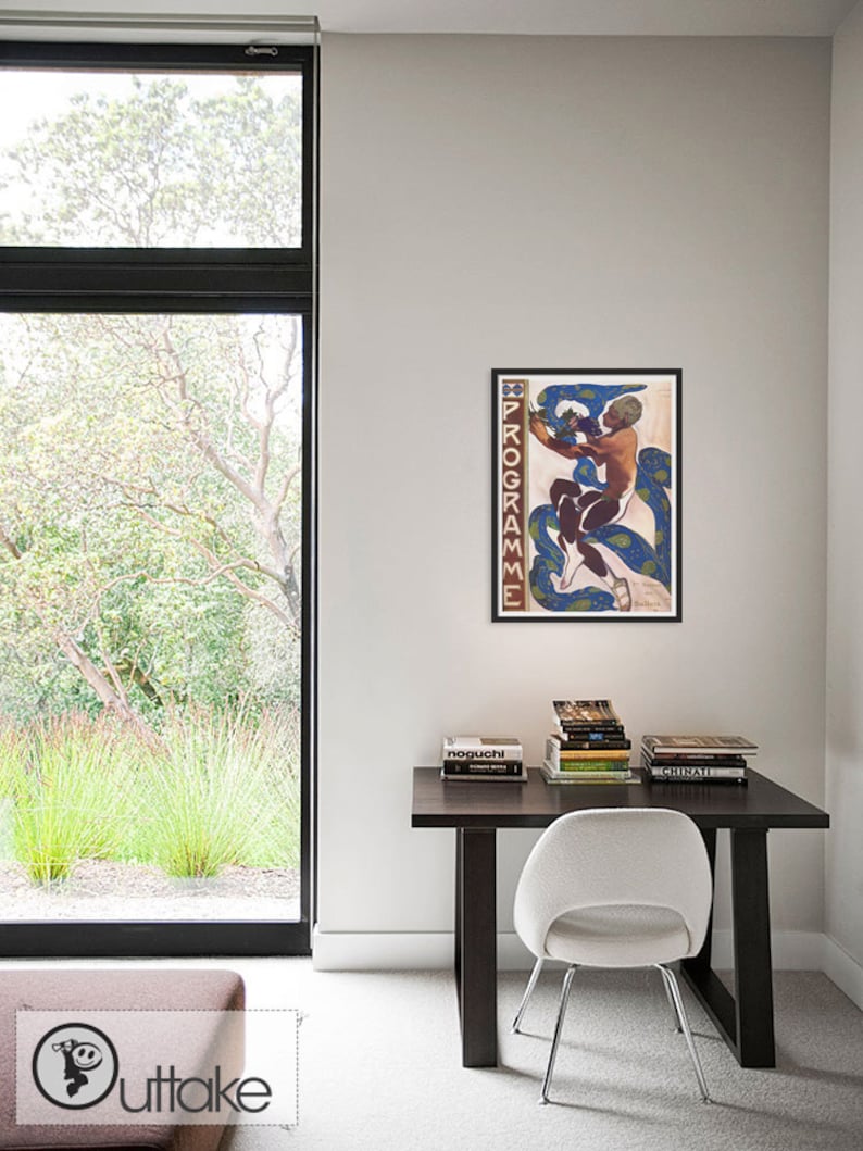 May include: A framed art print featuring a nude figure with blue and white serpentine forms. The artwork is displayed on a white wall above a dark wood desk with books and a white chair. The room has a large window with a view of greenery.