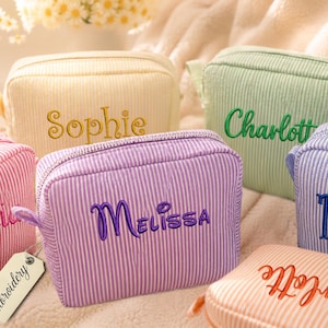 May include: A collection of rectangular, zippered cosmetic pouches made of striped fabric in pastel colors including yellow, green, purple, pink, and orange. Each pouch features custom embroidered names such as Sophie, Charlotte, and Melissa.