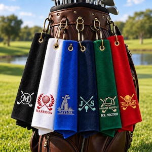 May include: Six rectangular golf towels in black, white, blue, navy, green, and red, each featuring a gold-toned metal grommet and a carabiner clip. Each towel displays unique embroidered golf-themed designs and names including M, Harrison, Taylor, J S, Mr. Victor, and Williams.