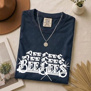 May include: A navy blue t-shirt featuring a white graphic print that reads "BEE GEES" in a stylized, angular font with starburst accents. The shirt has a Comfort Colors brand label at the inner neckline and is paired with a silver chain necklace.