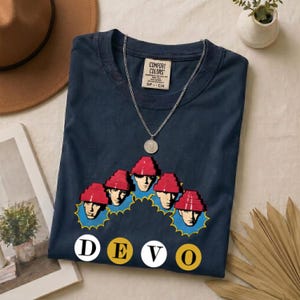 May include: A navy blue t-shirt featuring a graphic of five figures wearing red energy dome hats, each framed by a blue starburst. Below the figures, the word DEVO is printed in white and yellow circles. A silver chain necklace rests on the shirt.