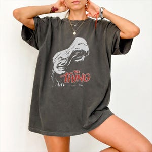 May include: A charcoal gray short-sleeve t-shirt featuring a distressed white graphic of a creature and the red text "THE THING" printed on the front. The shirt has a crew neckline and a relaxed, loose-fitting silhouette with a vintage-style wash.