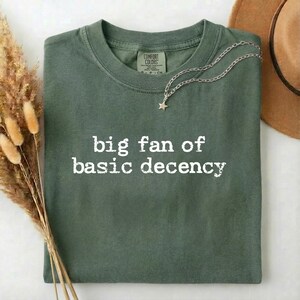 May include: An olive green t-shirt with the phrase "big fan of basic decency" printed in white. A silver necklace with a star pendant and a brown hat are also visible.