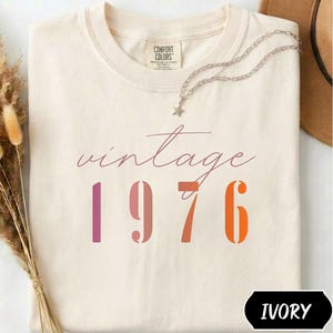 Könnte beinhalten: Ein elfenbeinfarbenes T-Shirt mit dem Text vintage 1976 in einer mehrfarbigen Schreib- und Blockschrift. Das Shirt hat einen Rundhalsausschnitt und ein Comfort Colors Markenetikett am Innenkragen. Eine silberfarbene Kette mit Sternanhänger liegt auf.