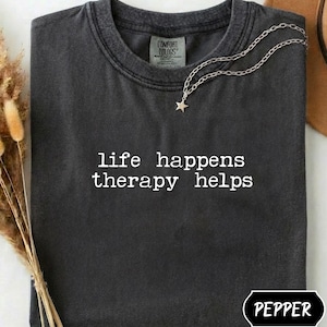 Könnte beinhalten: Dunkelgraues T-Shirt mit dem weißen Text "life happens therapy helps". Eine silberne Halskette mit Sternanhänger liegt auf dem Shirt. Das Shirt ist gefaltet und hat ein "Comfort Colors"-Etikett.