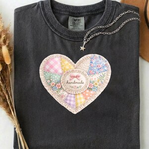 以下が含まれることがあります： パッチワークのハートのデザインが施された、濃いグレーのComfort Colors Tシャツ。ハートは様々なパステルカラーの生地でできており、中央には「handmade」の文字。星のペンダントが付いたシルバーのネックレスがTシャツの上にかけられています。