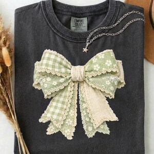 Op de afbeelding: Donkergrijs T-shirt met een groot strikdesign. De strik heeft een ruitpatroon, bloemenprint en kanten rand in groen- en crèmetinten. Een zilveren ketting met een sterhanger is over het shirt gedrapeerd.